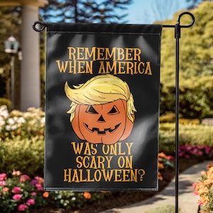 Puede incluir: Bandera de jard&iacute;n negra de Halloween con el texto "REMEMBER WHEN AMERICA WAS ONLY SCARY ON HALLOWEEN?" y una calabaza con pelo rubio. La bandera est&aacute; colgada de un poste de metal negro en un jard&iacute;n.
