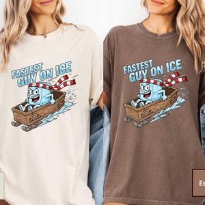May include: Ivory and espresso-coloured t-shirts with a cartoon ice cube character in a wooden sled. The ice cube wears a red and white striped scarf and the text "FASTEST GUY ON ICE" is above the character.