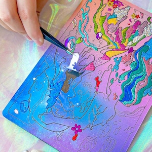 May include: A colorful art project in progress, featuring a vibrant illustration with blues, pinks, and greens. Tweezers are used to apply a small, white sticker to the artwork. Seashells and pearls are in the background.