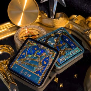 May include: Two square, teal-colored zodiac sign pins with gold accents and black borders. One pin features the Leo constellation, the other the Aquarius constellation. A compass and other gold-colored objects are in the background.