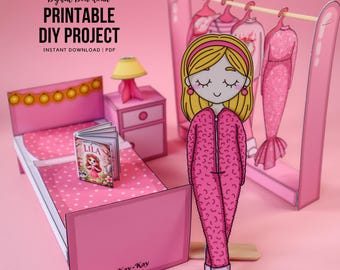 Peut inclure: Un ensemble de maison de poupée en papier rose avec un lit, une table de chevet et un portant. L'ensemble comprend une poupée en papier en combinaison rose. Le lit a une couette à pois roses et un livre intitulé "Lila". Le texte "Digital Download Printable DIY Project" est en haut.