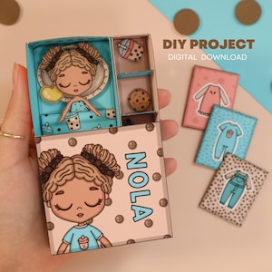 May include: A DIY project box with a doll illustration, featuring the name "NOLA". The box contains a doll, a cookie, and a drink. The box is held open, revealing the contents. Additional small cards with clothing illustrations are also present.