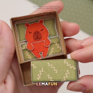 Pu&ograve; includere: Una scatola di cartone marrone in miniatura con un'illustrazione di un orso arancione addormentato all'interno. L'orso riposa su un cuscino e una coperta verdi. La scatola ha un cassetto con un motivo a foglie verdi. Il testo "LEMAFUN" &egrave; visibile sul cassetto.