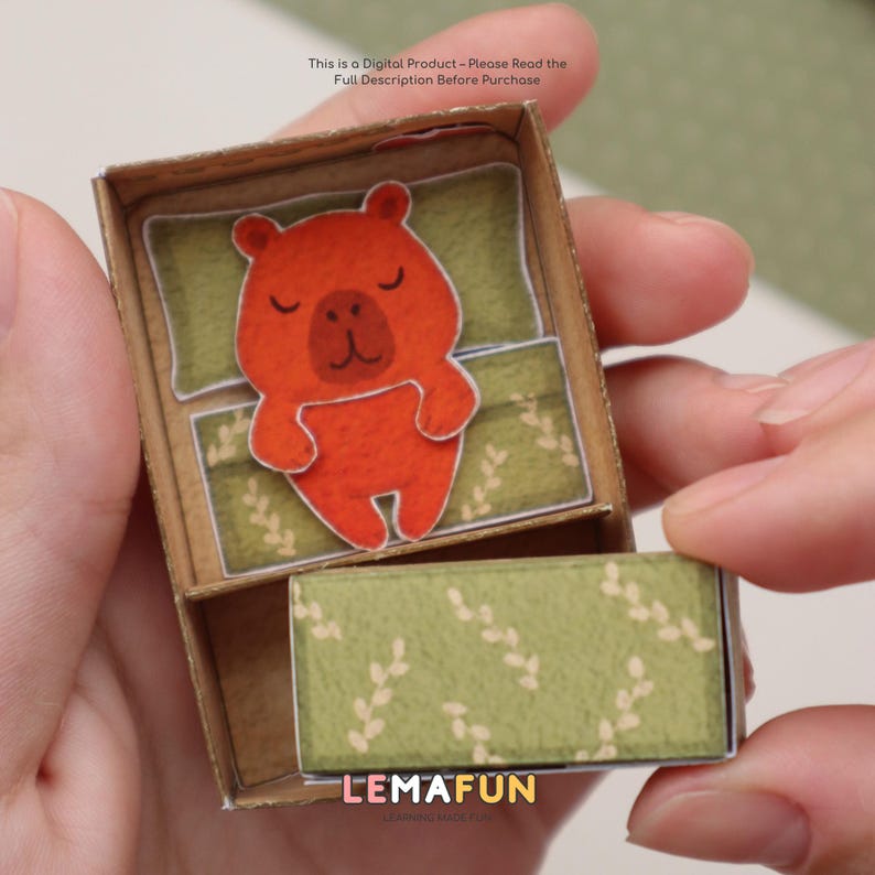 Pu&ograve; includere: Una scatola di cartone marrone in miniatura con un'illustrazione di un orso arancione addormentato all'interno. L'orso riposa su un cuscino e una coperta verdi. La scatola ha un cassetto con un motivo a foglie verdi. Il testo "LEMAFUN" &egrave; visibile sul cassetto.