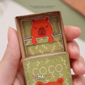 Pu&ograve; includere: Una scatola di carta artigianale con un orso arancione addormentato su un letto verde con un motivo floreale. La scatola &egrave; etichettata "COCO" con "LEMAFUN" e "LEARNING MADE FUN" sotto. Il testo in alto recita "This is a Digital Product - Please Read the Full Description Before Purchase."