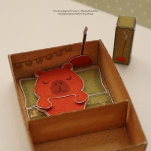 Pu&ograve; includere: Un diorama di carta con un orso arancione addormentato in un letto marrone con un cuscino verde. Sono presenti anche una piccola lampada e un comodino. Il testo "This is a Digital Product - Please Read the Full Description Before Purchase" &egrave; in alto.