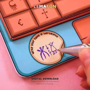 May include: A close-up of a colorful printable game board with the text "LEMAFUN" and "write your name in hieroglyphs". The game board is on a pink surface and a hand is holding a pen over a circle with hieroglyphs drawn inside.