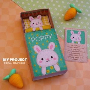 May include: A handmade DIY project featuring a matchbox with a bunny design. The box is green with the name "POPPY" and a bunny illustration. A small card with a bunny and text is next to the box. Two orange carrots with green tops are also present.