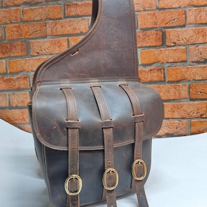 Può includere: Borsa a sella in pelle marrone con chiusura a patta e tre cinghie fissate con fibbie in ottone. La borsa ha una tracolla ed è progettata per essere indossata a spalla. La pelle ha una finitura invecchiata.