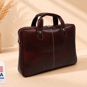 May include: A dark burgundy briefcase with a textured finish, two top handles, and a central vertical seam with white stitching. The bag features a top zipper closure and a white rectangular tag with the text USA DUTY FREE and a US flag.
