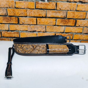 Puede incluir: Un cinturón de cuero negro con un diseño floral labrado en bronceado y marrón. El cinturón tiene una hebilla plateada y una correa de cuero a juego con una punta plateada. El cinturón es probablemente para una guitarra u otro instrumento.