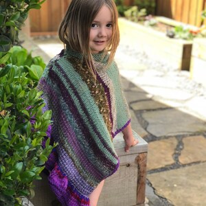 Op de afbeelding: Een kleurrijke, handgebreide poncho in groen, paars en bruin. De poncho heeft een hoge kraag en een gestreept patroon. De randen zijn afgewerkt met een levendige paarse rand. Het kledingstuk wordt door een jong kind gedragen.