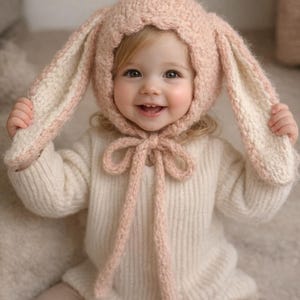 May include: A smiling toddler wearing a light pink and white bunny ear bonnet and a cream-colored sweater. The bonnet has long, floppy ears and a tie under the chin. The child is also wearing light pink pants.