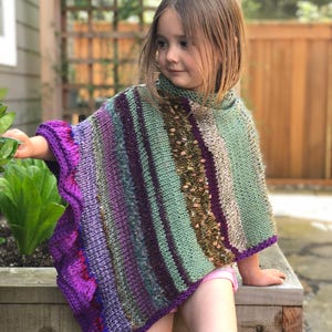 以下が含まれることがあります： 紫、緑、茶色の色合いの手編みのカラフルなポンチョ。 ポンチョは、スカラップエッジのテクスチャデザインが特徴です。 この衣服は幼い子供が着用しています。