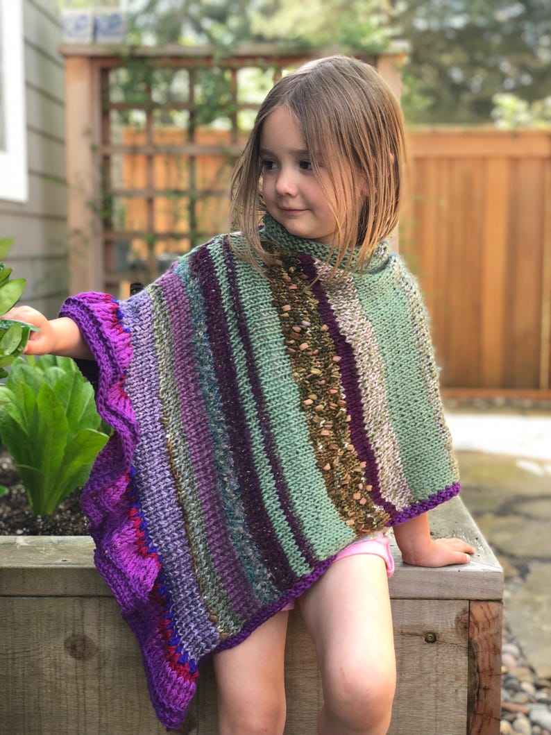 Op de afbeelding: Een kleurrijke, handgebreide poncho in paars, groen en bruin. De poncho heeft een textuurontwerp met een geschulpte rand. Het kledingstuk wordt door een jong kind gedragen.