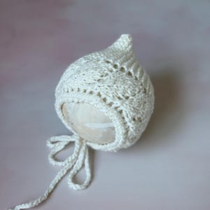 Könnte beinhalten: Ein weißer, gehäkelter Babyhut mit spitzer Spitze und filigranem Lochmuster. Der Hut hat einen Kordelverschluss und wird auf einem neutralfarbenen Requisit vor einem weichen, rosa Hintergrund präsentiert. Ein klassisches Baby-Accessoire.