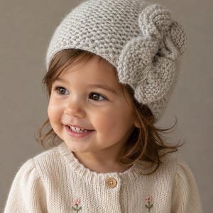 May include: A light grey knit beanie featuring a prominent, textured bow detail on the side. The hat is crafted with a dense, uniform stitch pattern and is paired with a cream-coloured, long-sleeved knit cardigan adorned with small floral embroidery.