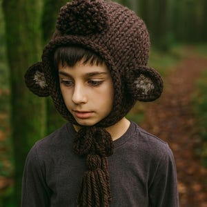 Può includere: Un cappello marrone lavorato a maglia con orecchie attaccate e una sciarpa con nappe. Il cappello ha un pompon sulla parte superiore ed è indossato da una persona. Lo sfondo è una scena di foresta sfocata.