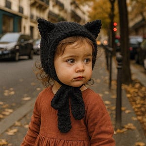 以下が含まれることがあります： 黒い猫耳ニット帽と、お揃いのスカーフを身につけた子供。子供はまた、長袖の錆色のドレスを着ています。背景には、車や木々のある街路が写っています。