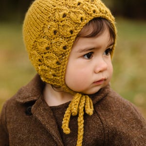 May include: Mustard yellow knitted baby bonnet with a textured floral pattern. The bonnet has a tie closure and is worn with a brown coat. The bonnet is a cozy accessory for infants and toddlers.