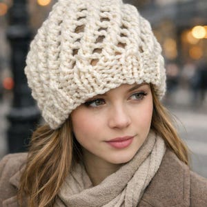 May include: Cream-colored, chunky knit hat with a textured design. The hat is worn with a matching scarf and a brown coat. The hat appears to be hand-knitted, offering a warm and cozy aesthetic.