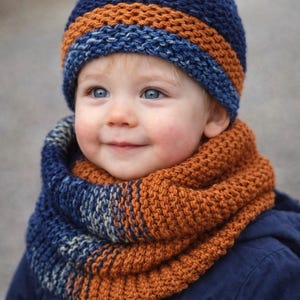 May include: A knitted hat and cowl set in shades of blue and orange. The hat has a blue base with an orange stripe, and the cowl features a similar color scheme. The set is made of a chunky knit material.