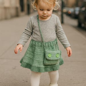 Op de afbeelding: Een jong kind draagt een grijze longsleeve, een groene gebreide rok, witte panty's en witte schoenen. Een kleine groene handtas met twee groene knopen hangt aan de schouder van het kind. De outfit is geschikt voor een peuter.