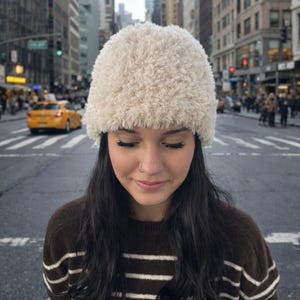 Könnte beinhalten: Ein flauschiger, cremefarbener Winterhut. Der Hut ist aus einem weichen, strukturierten Material gefertigt. Die Person trägt einen braun-weiß gestreiften Pullover. Der Hintergrund zeigt eine Stadtstraße mit Gebäuden und einem gelben Taxi.