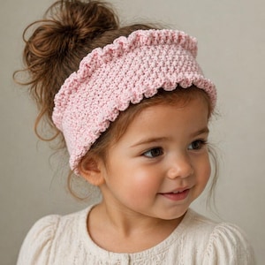 May include: A pale pink, crocheted headband with a scalloped trim. The headband is worn by a young child with brown hair. The child is wearing a cream-coloured button-down shirt. The background is a plain, neutral colour.