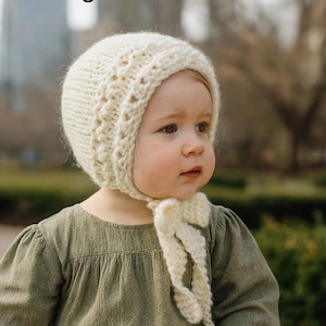 May include: A cream-colored knitted bonnet with a textured design and tie closure. The bonnet is worn by a child wearing a green dress. The words "Knitting Pattern" are displayed at the top of the image.