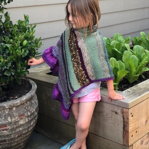 Op de afbeelding: Gebreide poncho voor kinderen in groen, paars en bruin. De poncho heeft een hoge kraag en een gestreept patroon. Het kind draagt een roze short en blauwe teenslippers.