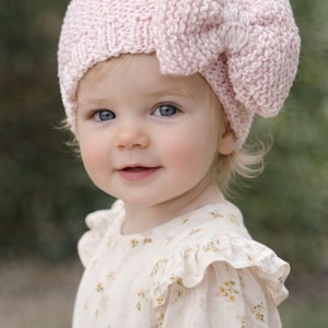 May include: A light pink knitted baby hat with a large bow detail. The hat is made of a chunky knit material. The baby is wearing a floral print dress.