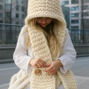 May include: A cream-colored, chunky knit hood and scarf set. The hood is oversized with a pointed top, and the scarf features a large wooden button closure. The set is made of a thick, textured yarn, perfect for cold weather.