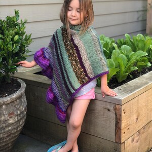 Op de afbeelding: Een handgebreide poncho in tinten paars, groen en bruin. De poncho heeft een hoge kraag en een gestreept patroon. Het kind draagt een roze short en blauwe teenslippers. De achtergrond omvat een houten plantenbak en groen.