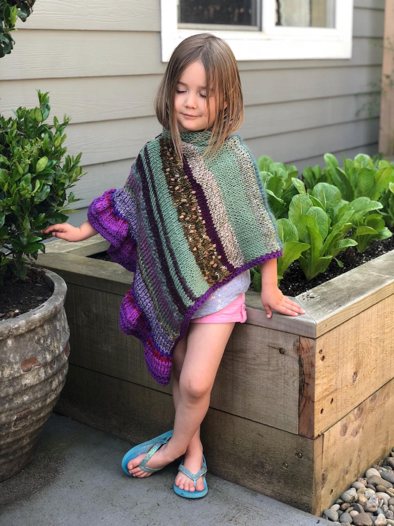 Op de afbeelding: Een handgebreide poncho in tinten paars, groen en bruin. De poncho heeft een hoge kraag en een gestreept patroon. Het kind draagt een roze short en blauwe teenslippers. De achtergrond omvat een houten plantenbak en groen.