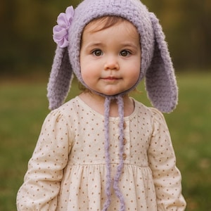 May include: A child wearing a lavender knitted bunny hat with long ears and a flower accent. The child is also wearing a beige dress with brown polka dots. The hat has ties under the chin.