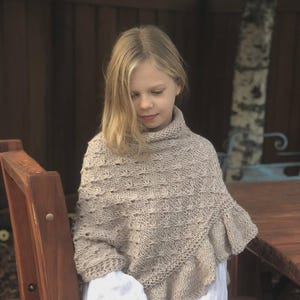 May include: A beige knitted poncho with a turtleneck and a ruffled hem. The poncho is worn over a white dress. The girl is standing outside near a wooden table and chair.