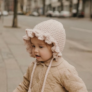 Könnte beinhalten: Ein hellrosa gehäkelter Babyhut mit gerüschtem Rand und geflochtenen Bändern. Der Hut wird von einem Kind getragen, das auch eine beige gesteppte Jacke trägt. Der Hintergrund ist eine verschwommene Szene im Freien.