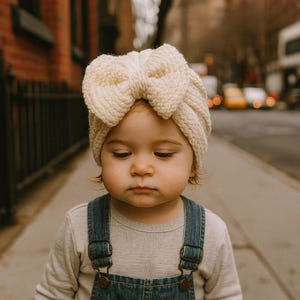 Può includere: Un bambino con una fascia a fiocco in maglia color crema, una maglietta a maniche lunghe grigio chiaro e una salopette di jeans. Il bambino è in piedi su un marciapiede davanti a un edificio in mattoni.