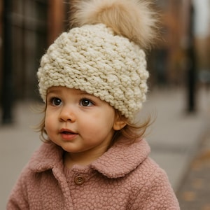 May include: A cream-colored, chunky knit hat with a fluffy, light brown pom-pom. The hat is worn by a young child, who is also wearing a pink, textured coat with a button closure. The image is taken outdoors.