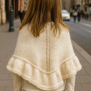 Può includere: Un mantello lavorato a maglia color crema con orlo a balze. Il mantello presenta un motivo centrale a trecce verticale e viene indossato sopra una camicia a maniche lunghe e pantaloni grigi. La persona è in piedi su un marciapiede.
