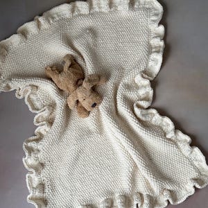 May include: A cream knitted baby blanket with a ruffled edge. A small brown stuffed animal is lying on top of the blanket.
