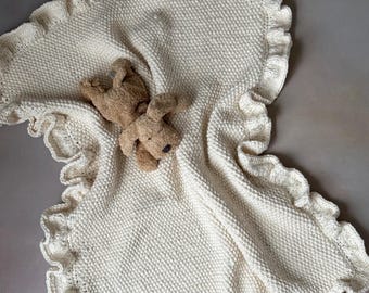 以下が含まれることがあります： クリーム色のフリル付きニットのベビーブランケット。ブランケットの上に小さな茶色のぬいぐるみが置かれています。