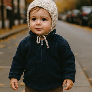 May include: A young child wearing a cream-colored knit hat with a pointed top and a navy blue fleece pullover with a half-zip closure. The child is also wearing brown corduroy pants. The background shows a street with trees.
