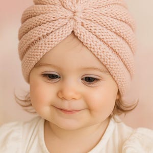 以下が含まれることがあります： 淡いピンク色のニットターバン風ベビーハット。柔らかく、質感のある糸でできており、上部にひねりのあるノットディテールが特徴です。赤ちゃんは白いシャツを着ています。