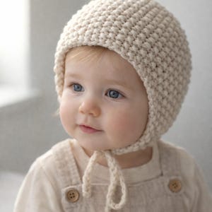 May include: A cream-colored, knitted baby bonnet with a rounded crown and chin ties. The bonnet is worn by a baby with blue eyes and fair skin, dressed in a cream-colored onesie. The overall aesthetic is soft and gentle.
