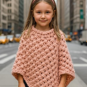 May include: A light pink, chunky knit poncho with a round neckline. The poncho is worn by a young person with long blonde hair and a pink headband. The background features a city street with tall buildings.