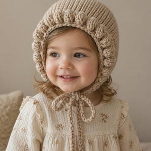 May include: A beige knitted bonnet with a scalloped edge and a tie closure. The bonnet is worn by a child wearing a cream-colored dress with a floral pattern. The background is a neutral beige.
