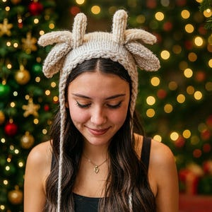 May include: A knitted beige reindeer antler hat with long braided ties. The hat features a headband and four upright antler shapes. The background includes a Christmas tree with ornaments and lights.