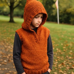 Può includere: Un gilet con cappuccio arancione lavorato a maglia con scollo a V e cappuccio a punta. Il gilet è realizzato con filato spesso e sembra caldo. La persona indossa sotto una camicia nera a maniche lunghe. Lo sfondo è un parco con alberi e foglie cadute.